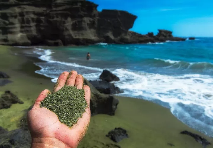 Mano con olivina molida en primer plano y al fondo una playa con arenas verdes. Project Vesta y Climitigation